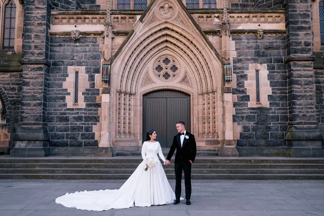 A bride and groom holding hands in front of a Gothic-style church facade.