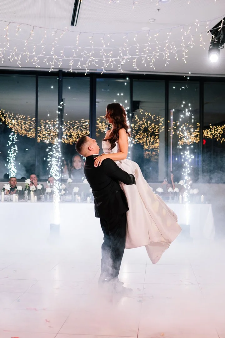 A couple dancing at their wedding reception, with the man lifting the woman in a ballroom decorated with string lights and fireworks.
