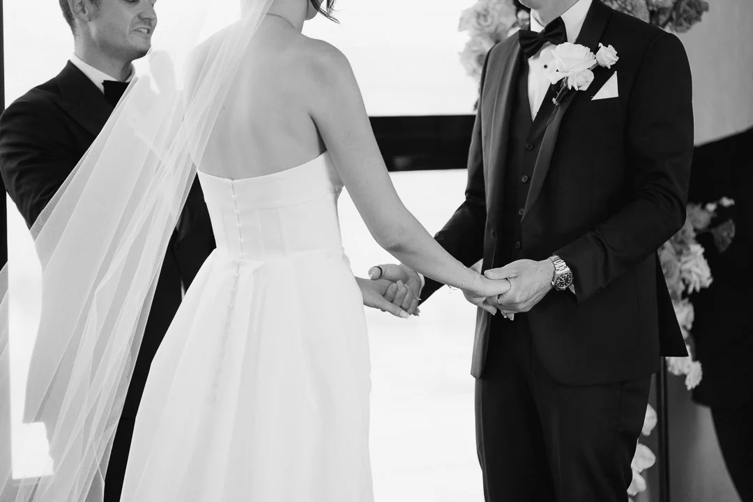 A wedding ceremony where a bride in a white strapless dress and a veil holds hands with a groom in a tuxedo, during their vows.