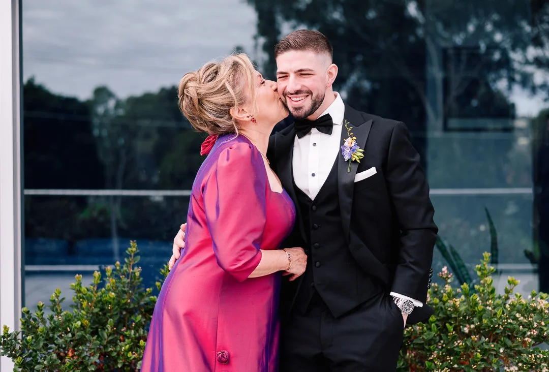 A woman in a pink and purple dress and a man in a black tuxedo with a bow tie, smiling and sharing a kiss on the cheek outdoors on a balcony with greenery.