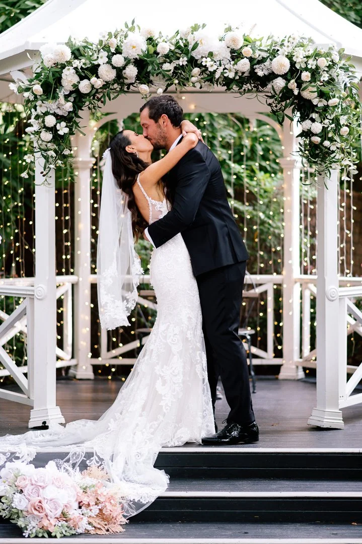 A bride and groom kissing underneath a floral wedding arch at their outdoor wedding ceremony, with fairy lights in the background.