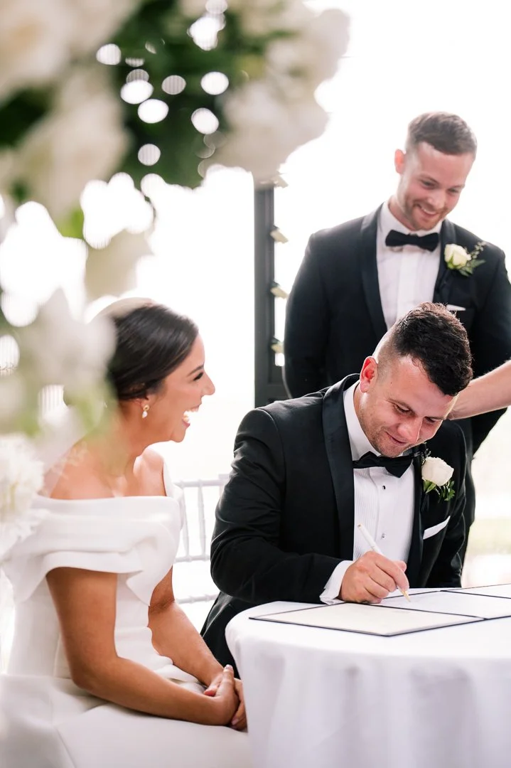 A bride and groom signing a marriage document during their wedding ceremony, with two groomsmen standing behind them. The bride is smiling, wearing a white off-the-shoulder gown, and the groom is signing with a pen, wearing a black tuxedo with a whit