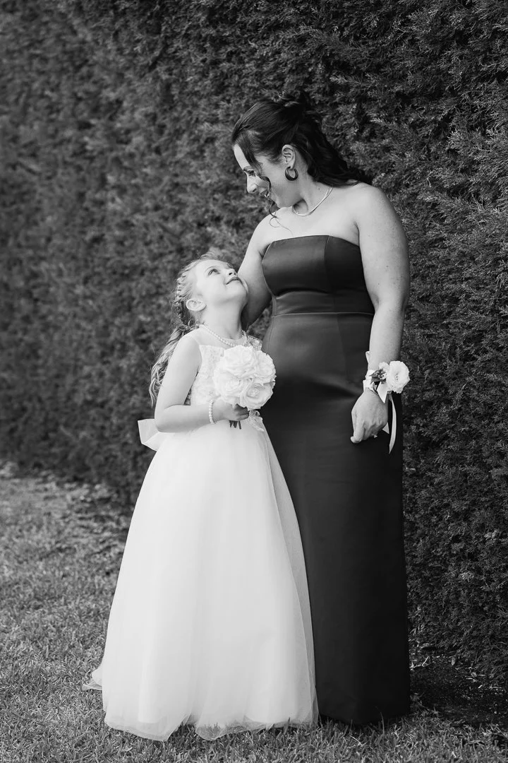 A woman in a strapless black evening gown and a young girl in a white princess dress holding a bouquet of flowers look at each other fondly outdoors with a hedge in the background.