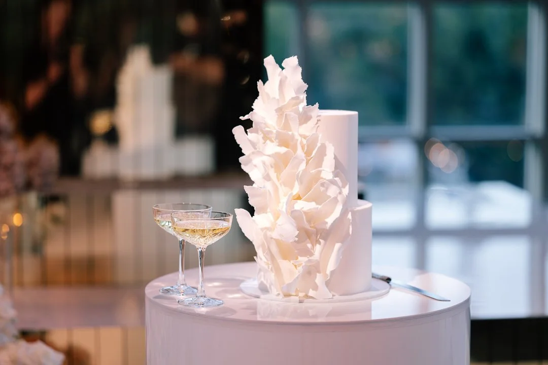 Elegant white wedding cake with floral decorations on a table, accompanied by two champagne glasses, in a bright indoor setting.