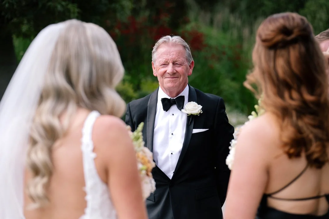 An elderly man in a tuxedo with a white flower boutonniere, smiling happily, looking at two women in wedding dresses during a wedding ceremony outdoors.