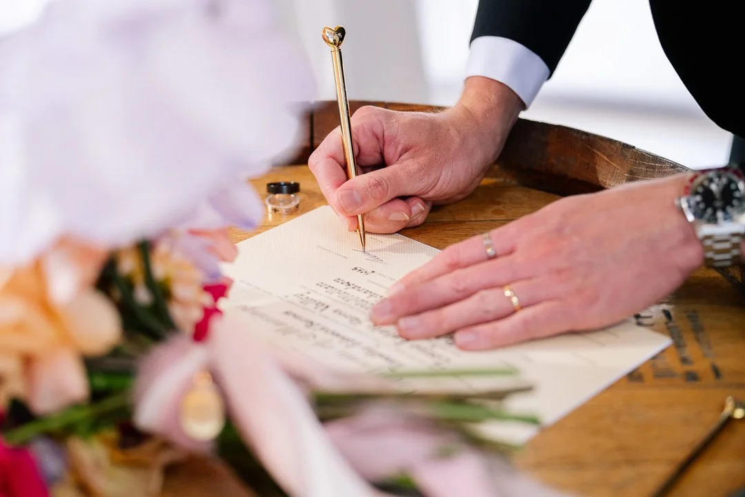 Close-up of hands of a person in a suit signing a document with a gold pen on a wooden table, with a bouquet of flowers nearby.