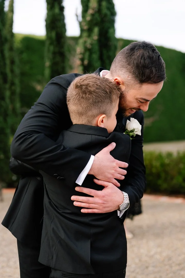 Two men in formal black suits embrace and smile outdoors, likely at a wedding or special event, with green trees in the background.
