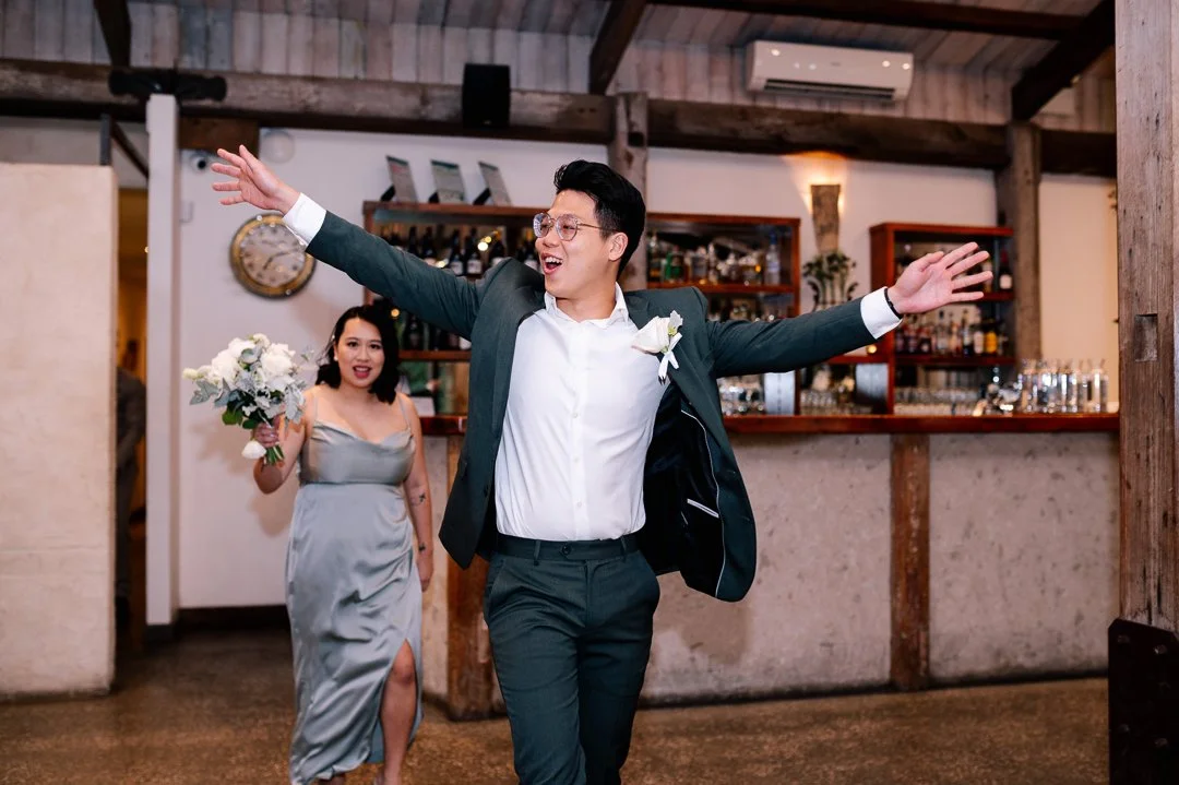 A man and a woman celebrating at a wedding reception, with the man dancing joyfully with arms outstretched and the woman holding a bouquet of flowers.