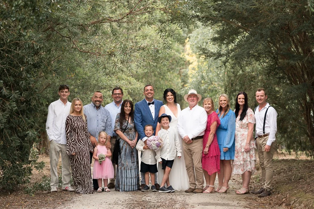 Family group photo outdoors among trees, celebrating a special occasion, with some members in formal attire and others in casual dresses, smiling.