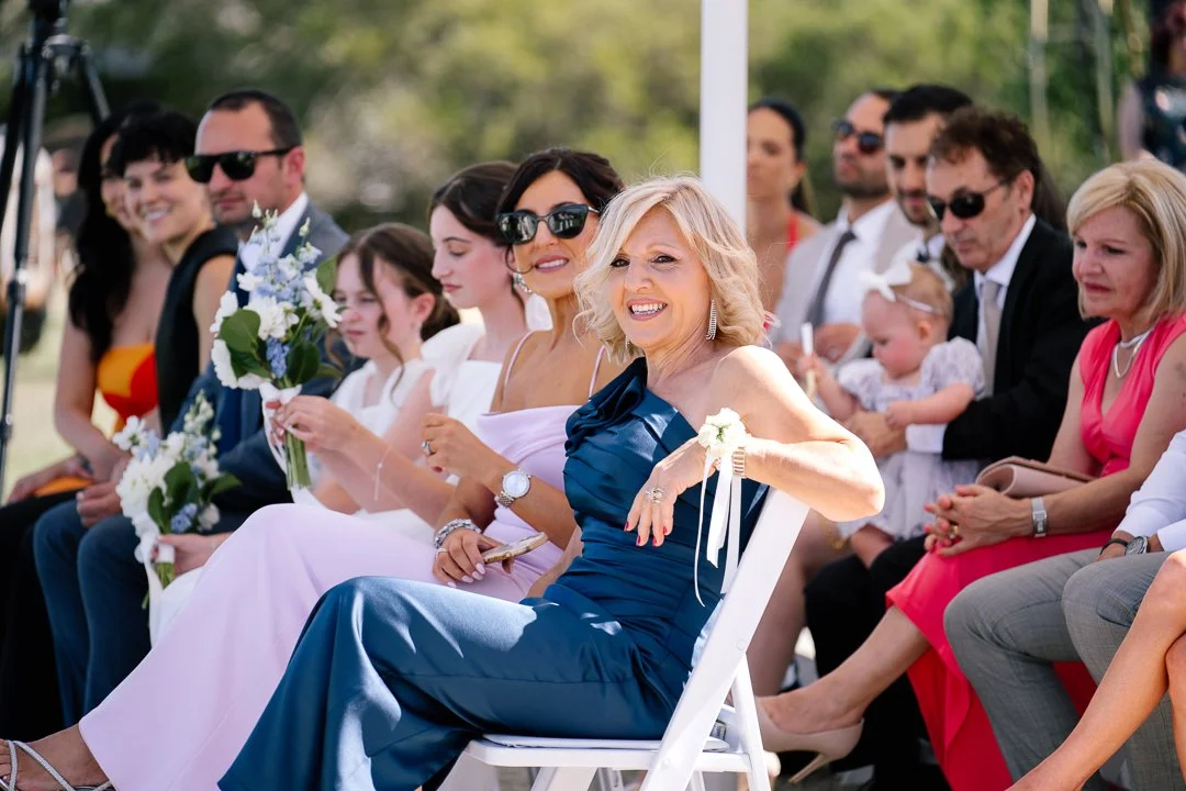 People attending an outdoor wedding ceremony, sitting on white chairs in formal attire, with some holding bouquets, and a woman in the front wearing a dark blue dress smiling.