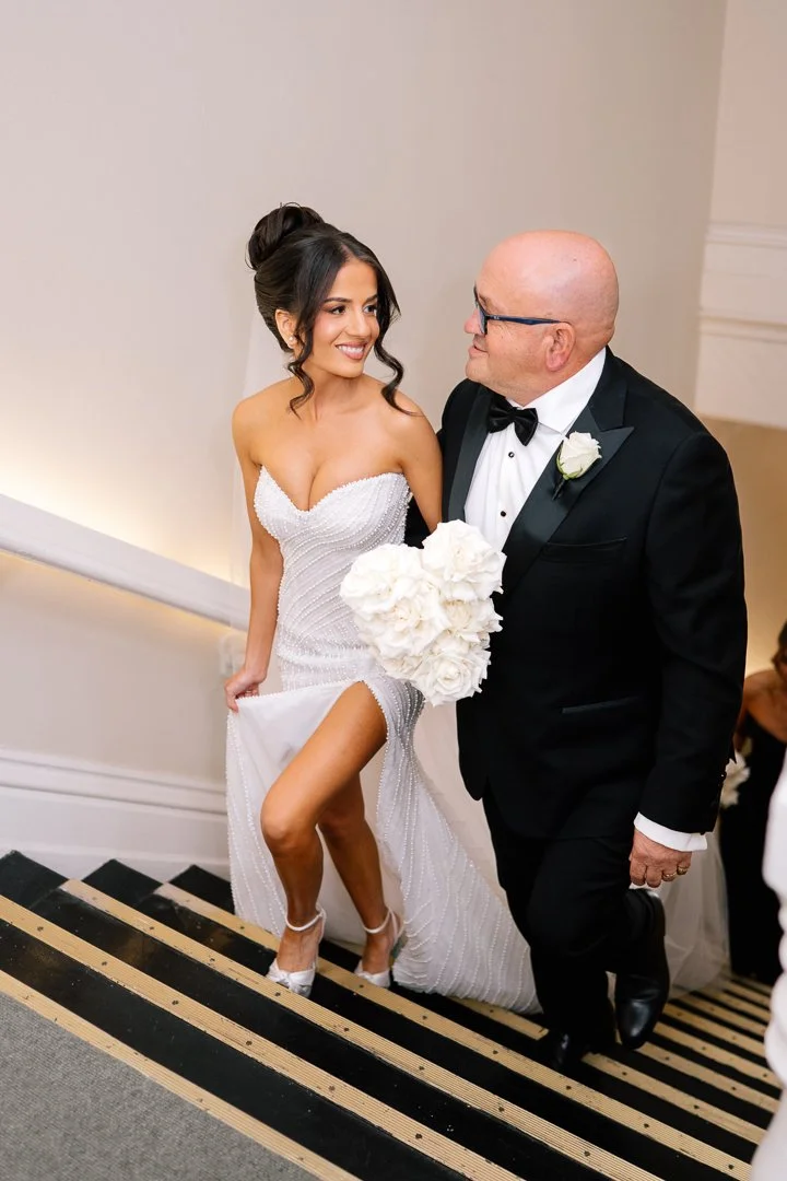 A bride in a white strapless wedding dress holding a bouquet of white flowers, smiling and looking at a man in a tuxedo, likely at a wedding celebration.