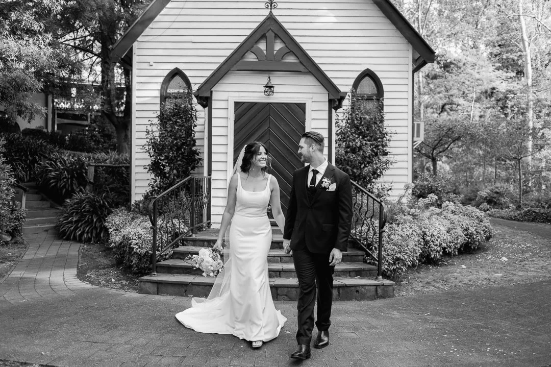 A bride and groom walk hand in hand outside a small church or chapel, with the bride holding a bouquet of flowers, surrounded by greenery and flowering bushes.