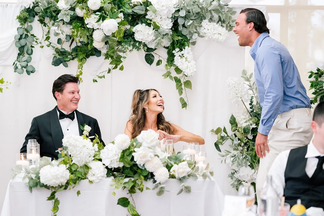 A wedding reception scene with a bride and groom seated at a table decorated with white flowers and candles, smiling and laughing, while a man standing and reaching out is also smiling.