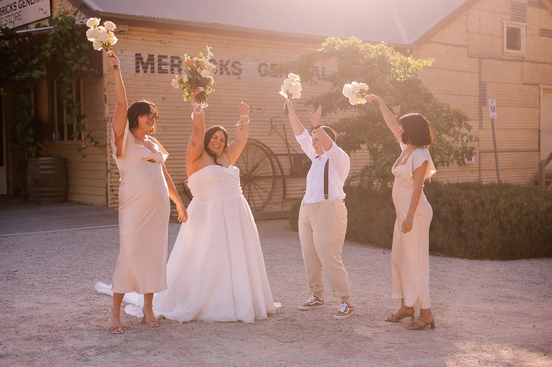 Four women celebrating outdoors at sunset, with two women holding bouquets, one in a wedding dress and others in dresses, raising their arms in joy.