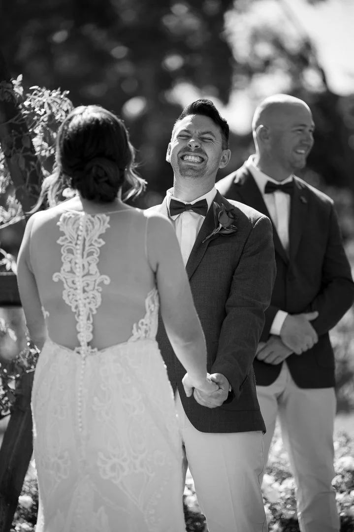 A black and white photo of a wedding ceremony with the bride and groom holding hands, exchanging vows, outdoors with trees in the background. The groom is smiling with his eyes closed, wearing a tuxedo with a bow tie and boutonniere. The bride's back