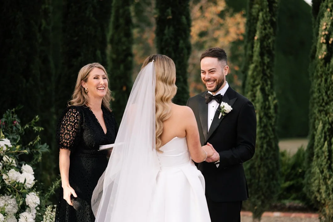 A bride and groom exchanging vows and holding hands during their outdoor wedding, with a woman officiant standing nearby in front of an arch of greenery and flowers.
