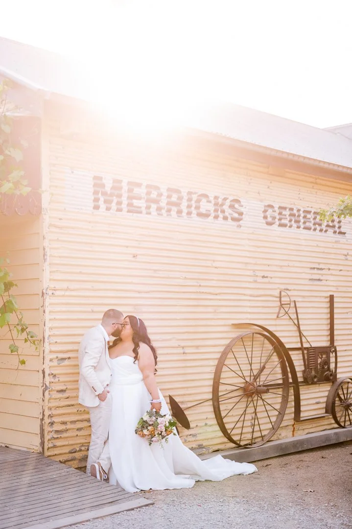 A newlywed couple in wedding attire sharing a kiss outside a rustic building with a sign that reads 'MERRICK'S GENERAL.' The bride holds a bouquet and the groom is in a light-colored suit.