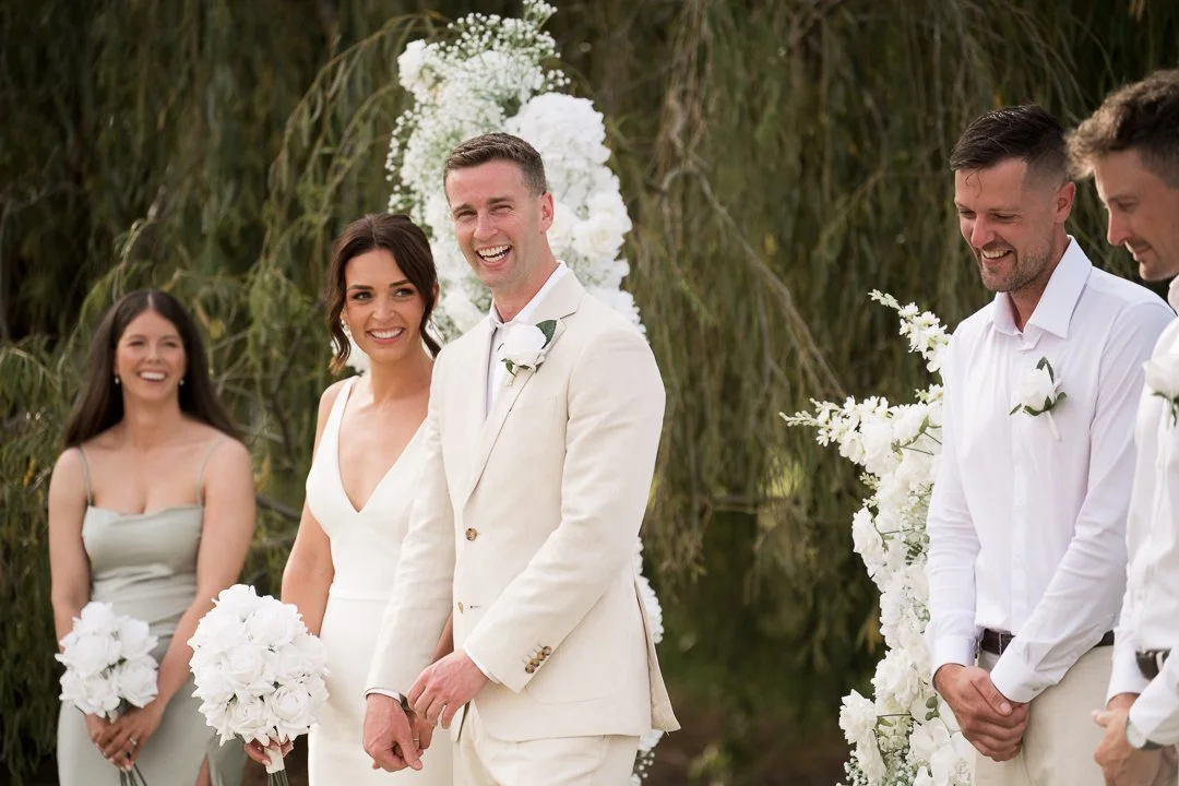 A wedding ceremony outdoors with a bride, groom, and three bridesmaids and groomsmen, all dressed in light-colored attire, with floral decorations around.