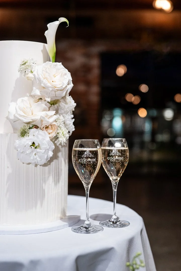 A wedding cake decorated with white flowers and two glasses of champagne on a white tablecloth, with a blurred background.