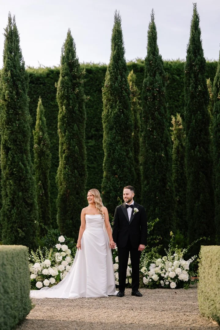 Bride and groom holding hands and smiling at a wedding ceremony outdoors, surrounded by tall green trees and white floral arrangements.
