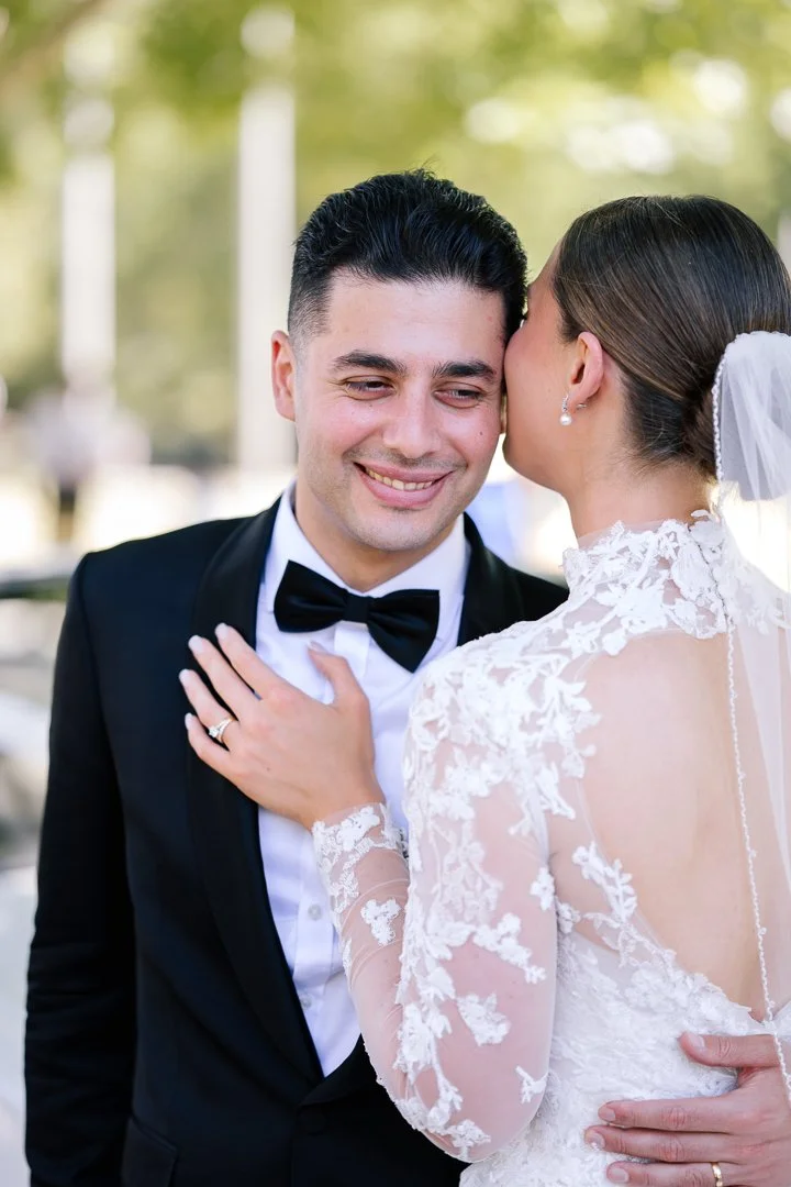 A bride whispering to a groom in a black tuxedo outdoors on a wedding day.