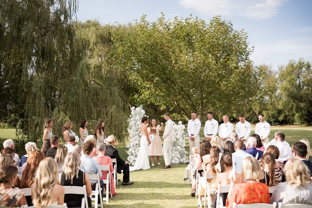 An outdoor wedding ceremony taking place in a park with a large green tree and a white floral arch as a backdrop. The bride and groom are standing before a officiant, surrounded by bridesmaids and groomsmen, with guests seated on white chairs watchin