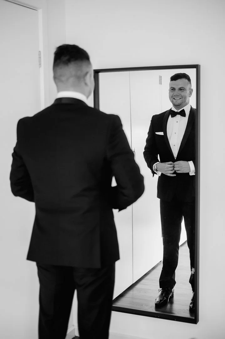 A man in a tuxedo looking at himself in a full-length mirror, adjusting his suit, with another man visible in the reflection.