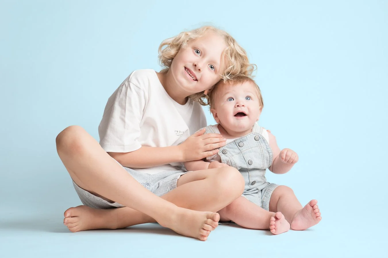 Two young children, one with curly blonde hair and the other with red hair, sitting on a light blue background. The older child is smiling and leaning towards the younger, who has a surprised or curious expression.