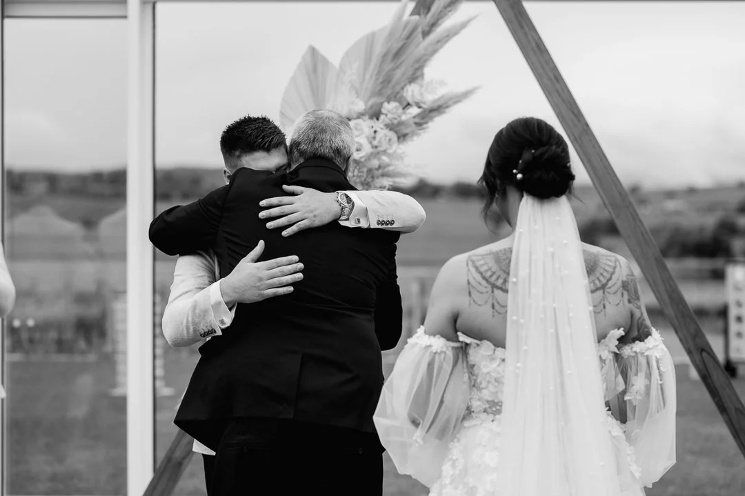 Two men embrace during a wedding ceremony, with a bride standing nearby under an arch decorated with flowers and pampas grass, outdoors with a field in the background.