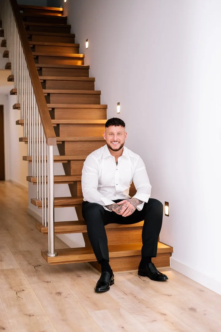 A smiling man with tattoos on his hands and neck, wearing a white dress shirt and black pants, sitting on a wooden staircase in a modern home.