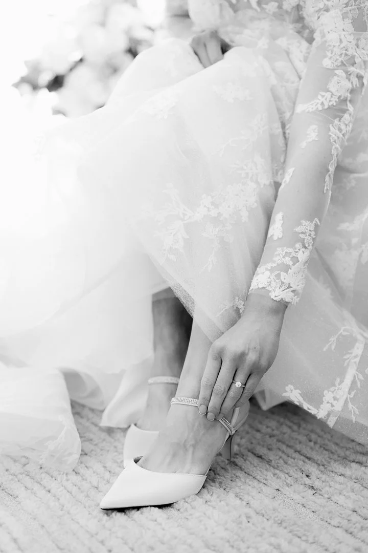 A bride adjusting her high-heeled shoe while sitting on a carpeted floor, wearing a lace wedding dress and jewelry.