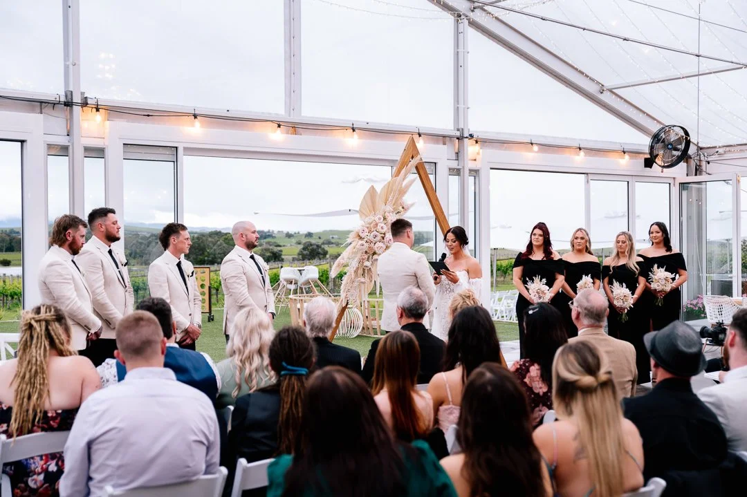 A wedding ceremony inside a glass venue with a bride and groom facing each other, officiating a wedding, in front of guests. The bride is in a white dress and the groom in a light-colored suit. Bridesmaids and groomsmen stand on either side, with the