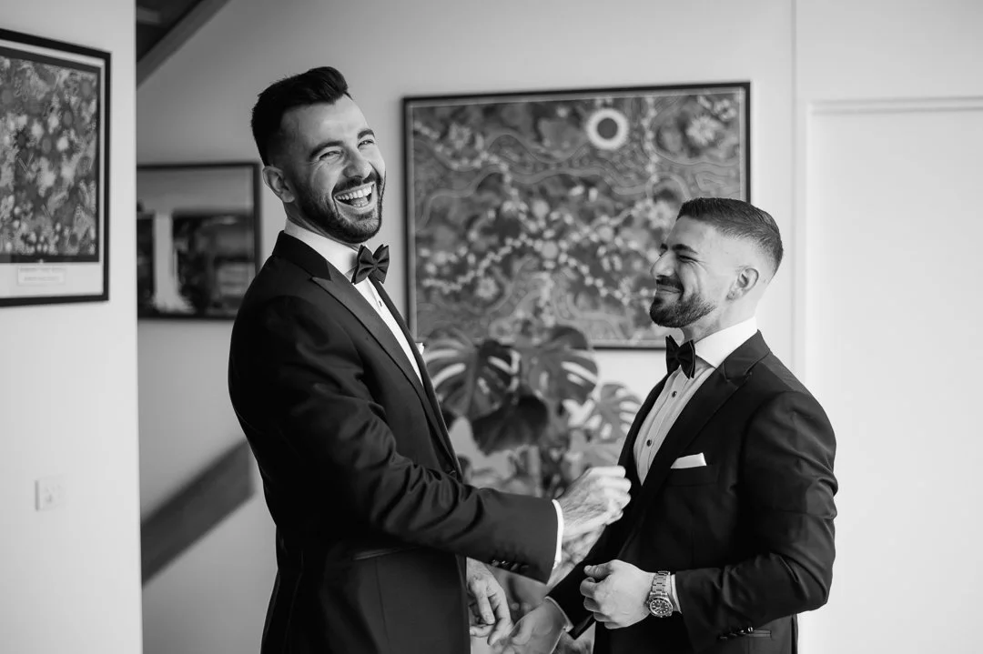 Two men in tuxedos sharing a laugh, holding hands indoors, with framed artwork and houseplants in the background.