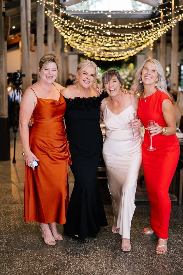 Four women in elegant dresses standing together at a celebration, smiling, with string lights hanging overhead in a rustic decorated venue.