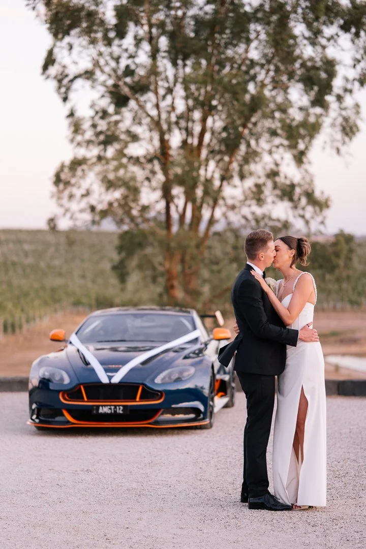 A bride and groom kiss in front of a decorated black sports car with white ribbons, set outdoors with trees and a field in the background.