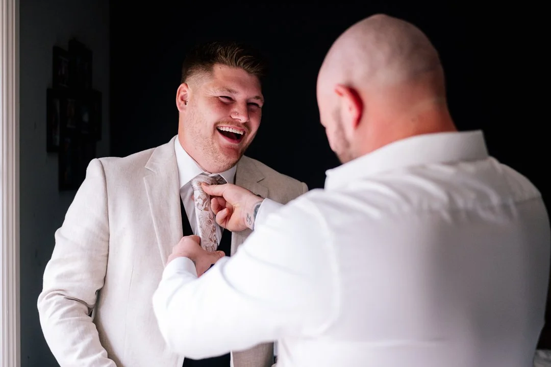 Two men in formal attire smiling and laughing, one helping the other with a tie.