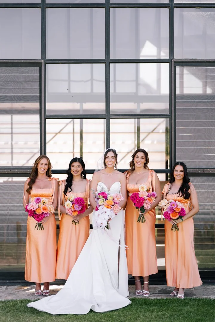 A bride in a white wedding gown holding a bouquet of white, pink, and orange flowers, standing between four bridesmaids in matching peach dresses, each holding a colorful bouquet of pink, orange, and white flowers, in front of a modern glass building