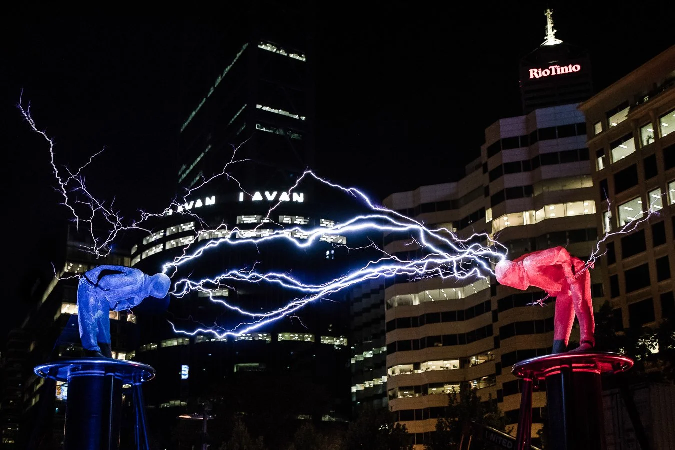 Night scene of two statues of people bowing on pedestals, connected by electric lightning bolts between their hands, set against a cityscape with illuminated buildings and the Rio Tinto sign.