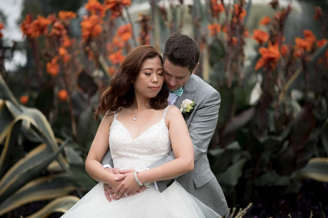 A couple dressed in wedding attire, standing outdoors in front of orange flowering plants. The bride is wearing a white wedding gown and the groom is in a gray suit. The groom is embracing the bride from behind, with his head tilted towards her.