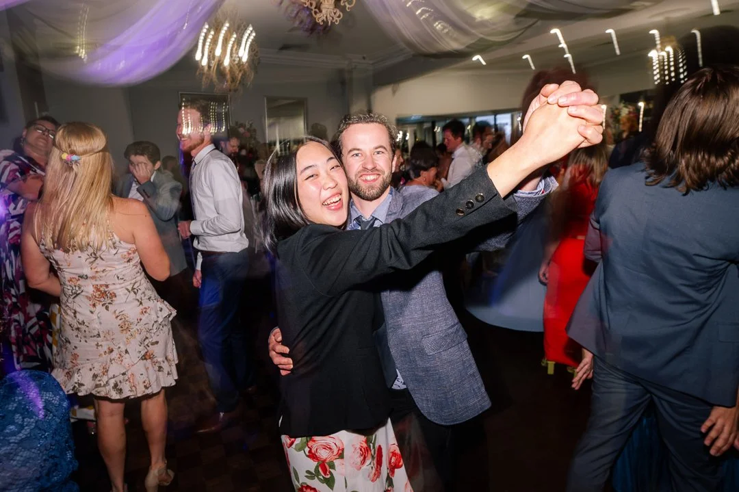 People dancing at a lively party or celebration in an indoor venue, with a couple twirling and smiling at the center of the image.