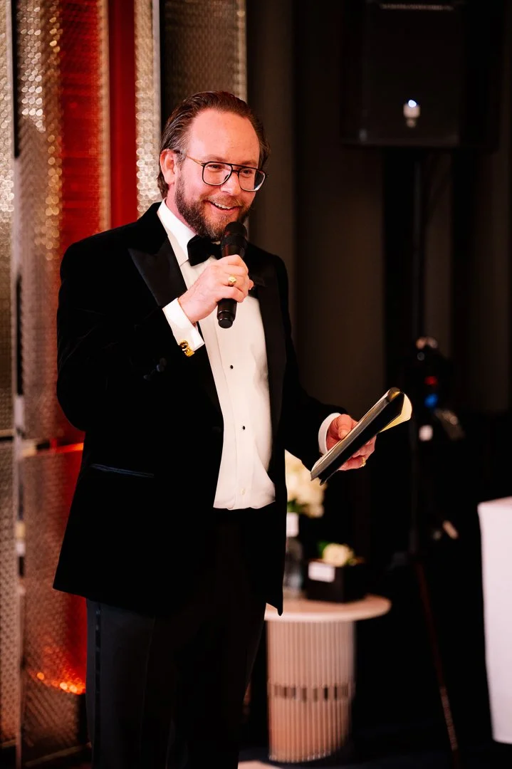 A man in a tuxedo with a bow tie, glasses, and a beard, holding a microphone and a notepad, speaking at an event.