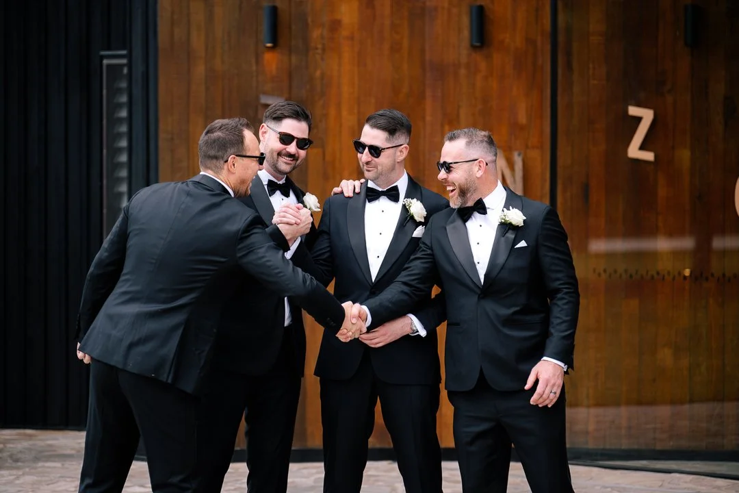 Four men in black tuxedos and sunglasses, with white corsages, shaking hands and smiling in front of a wooden wall.