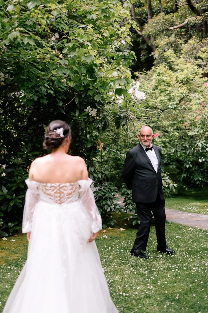 A bride in a white wedding dress standing with her back to a man in a black tuxedo in a lush garden.
