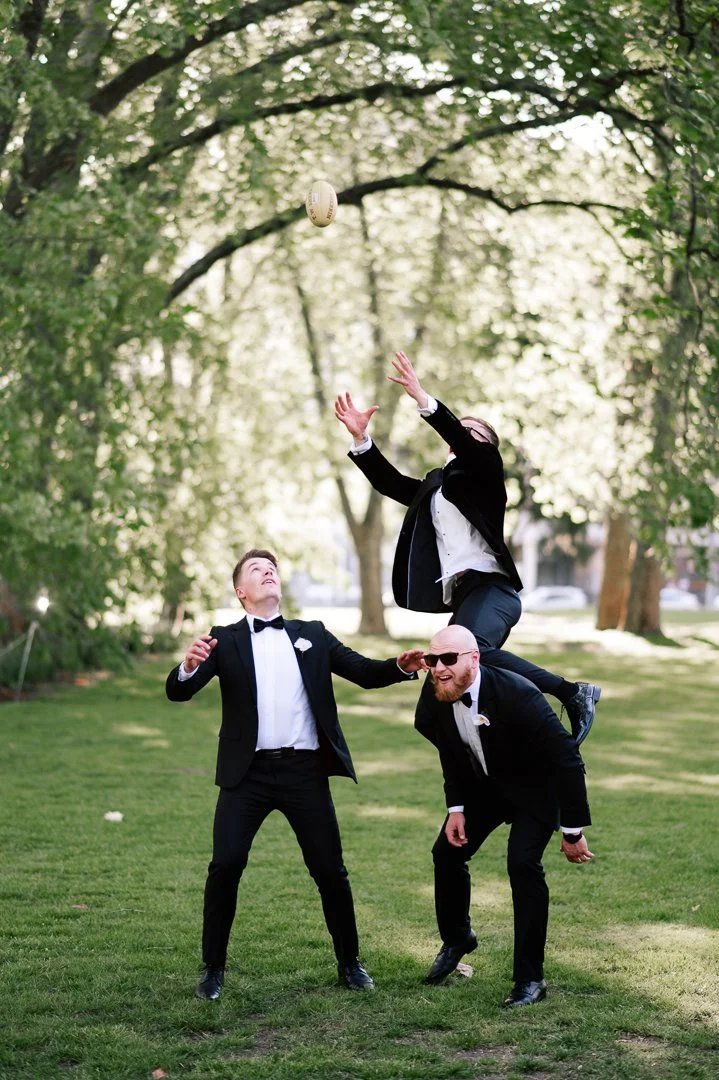 Three men dressed in tuxedos playing with a rugby ball outdoors in a park with trees.