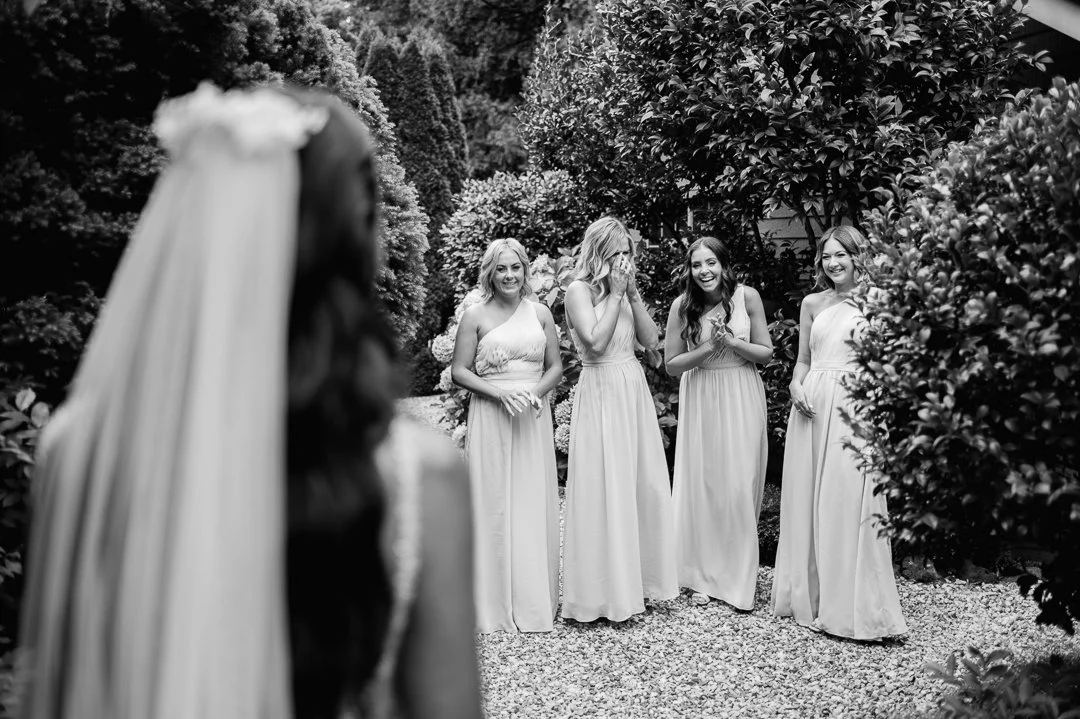 A bride with a veil looks at four bridesmaids in a garden, all wearing long dresses, with one covering her face with her hands in surprise or emotion.