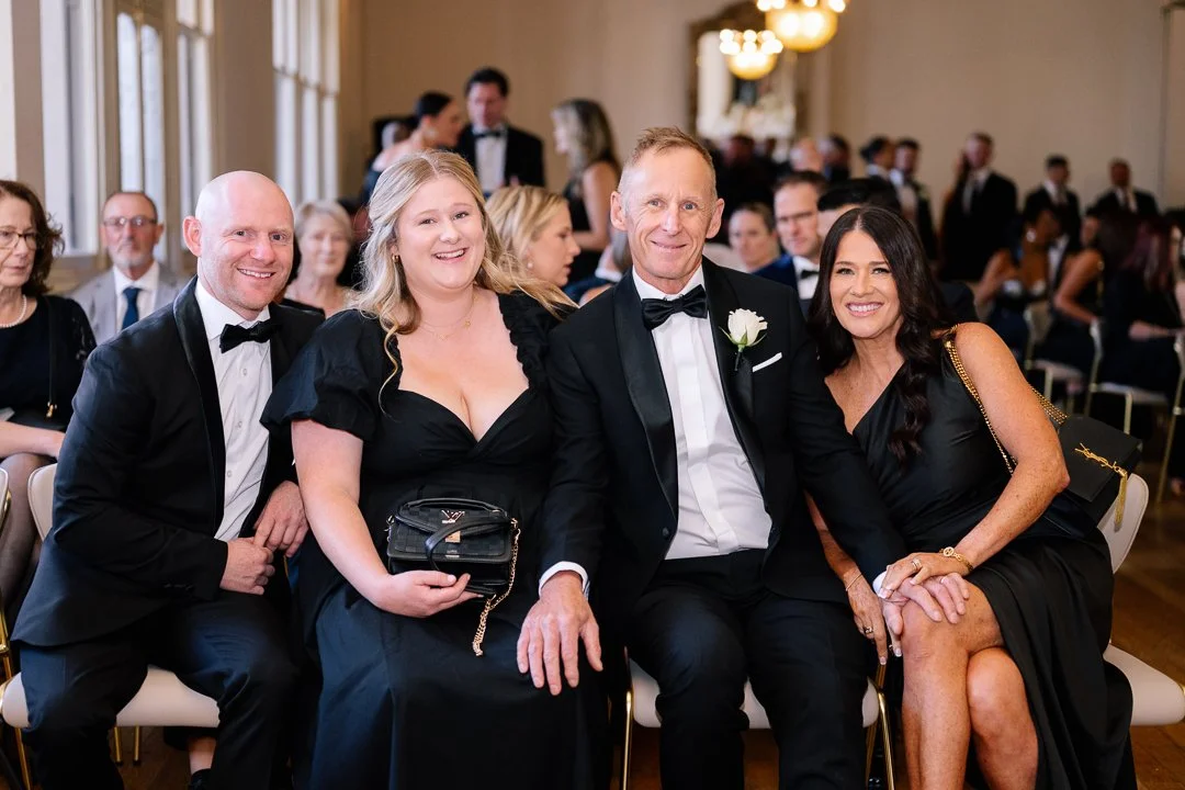 Group of four people in formal attire sitting together at an event, smiling and posing for a photo, with a room full of other guests in the background.