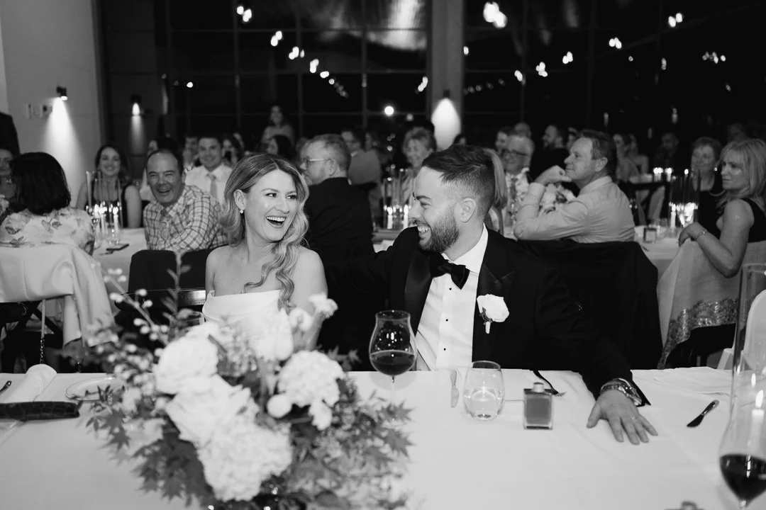 A bride and groom smiling and laughing at their wedding reception, seated at a table with a floral centerpiece, guests in the background, and string lights overhead.