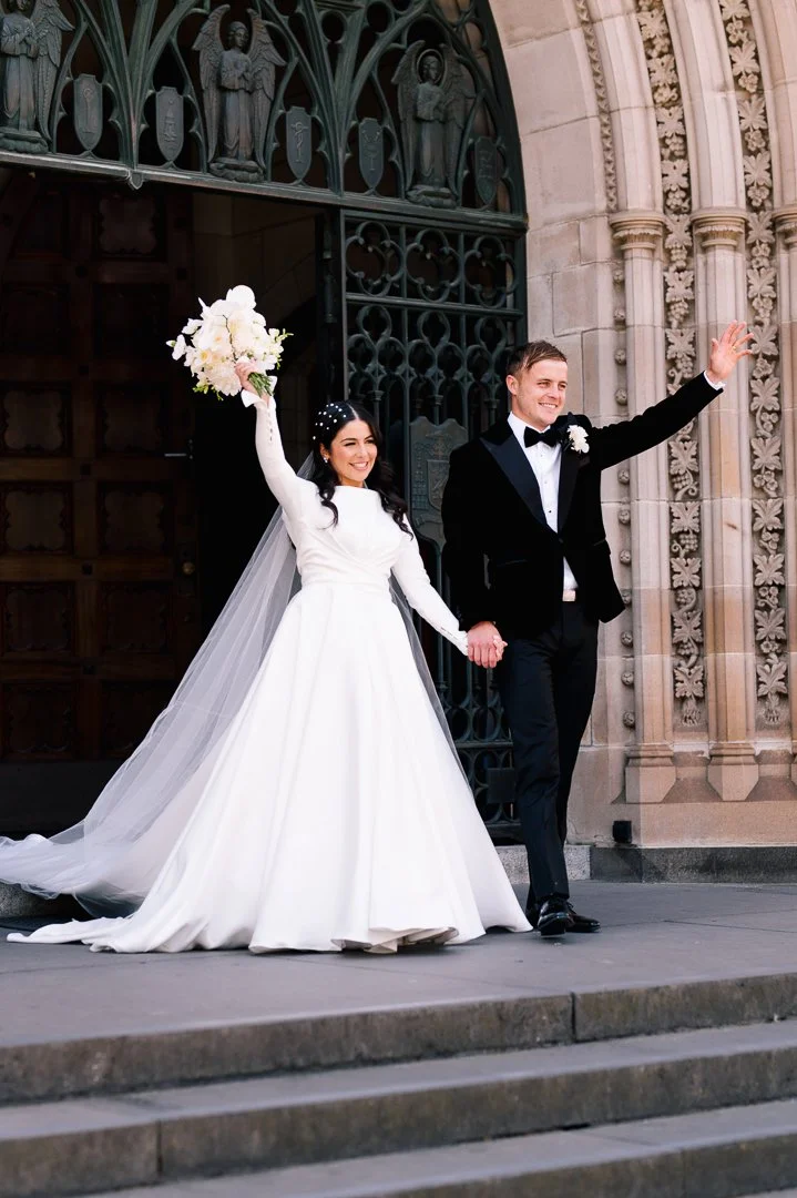 Bride and groom holding hands, smiling, and waving outside a church, wedding day.