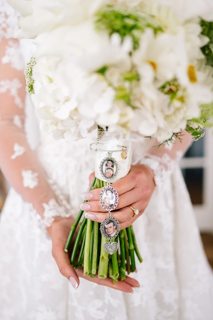 Bride holding a bouquet of white flowers with small photo charms hanging from the stems, wearing a white lace dress and a gold ring.
