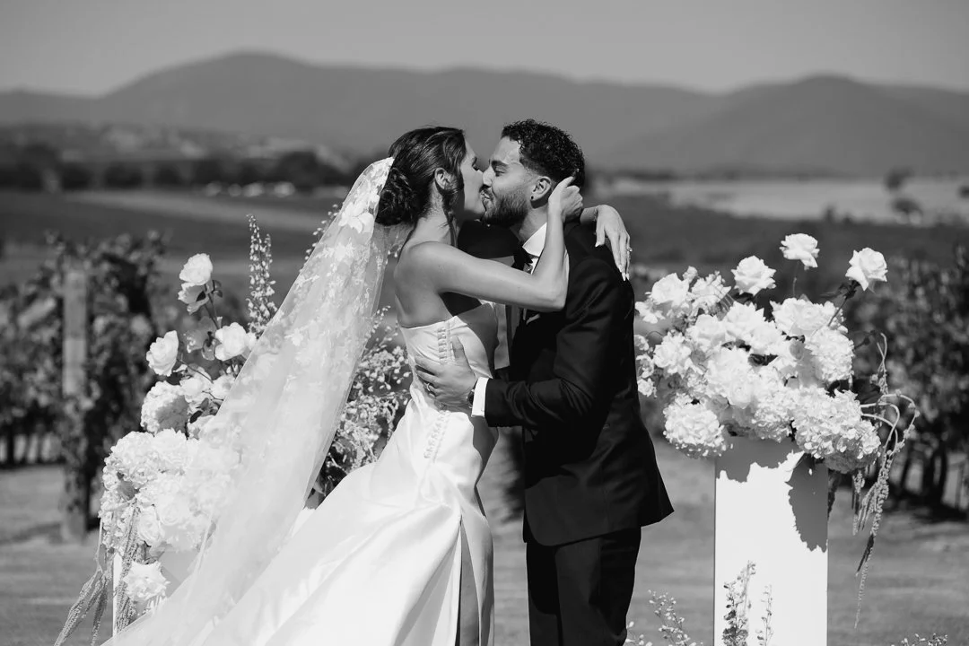 A couple in wedding attire embracing outdoors, with the bride in a white gown and veil, and the groom in a dark suit, surrounded by large flowers with hills in the background.
